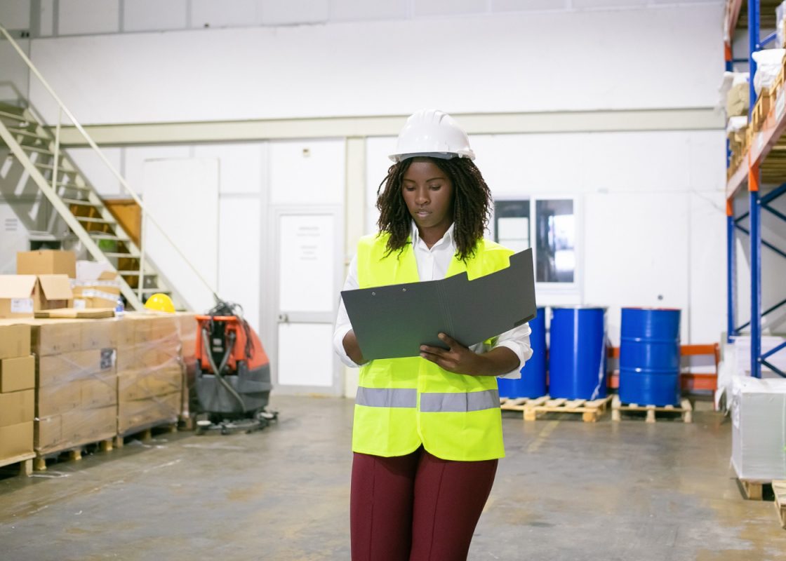 Focused Female Logistic Employee Hardhat Safety Vest Walking Warehouse Carrying Open Folder Looking Through Document Copy Space Front View Labor Inspection Concept (1)