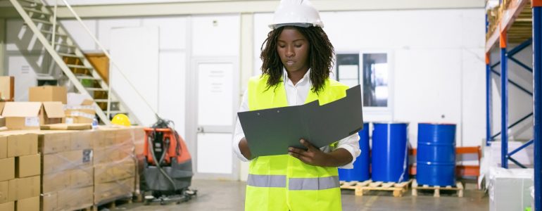 Focused Female Logistic Employee Hardhat Safety Vest Walking Warehouse Carrying Open Folder Looking Through Document Copy Space Front View Labor Inspection Concept